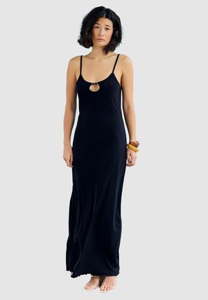 Femme aux cheveux noirs ondulés portant une longue robe noire sans manches avec une petite découpe sur la poitrine et des bracelets en bois à son poignet droit.