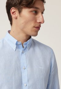 Chemise boutonnée en lin bleu clair avec une texture douce, un col pointu et un petit logo de chien brodé sur la poitrine.