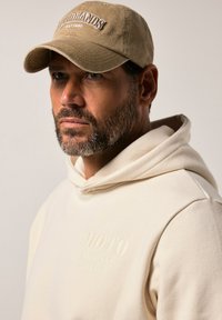 Felpa leggera beige con tessuto testurizzato e tasca a marsupio, con logo in rilievo "MOTO COMPANY JP 1880". Cappellino beige con testo "WOODLANDS EST. 1880".