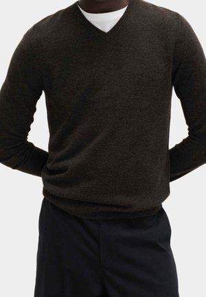 Mand iført mørkebrun V-hals sweater over hvid T-shirt, kombineret med sorte bukser mod en ensfarvet lys baggrund.