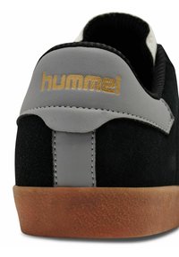 Zwarte suède sneaker met grijze leren accenten, voorzien van een gouden "hummel" logo, een gestructureerde rubberen zool en contrasterende stiksels.