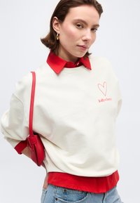 Νεαρή γυναίκα που φοράει λευκό φούτερ με κόκκινο λογότυπο καρδιάς, κόκκινο πουκάμισο με γιακά από κάτω, κόκκινη τσάντα ώμου και μπλε τζιν.