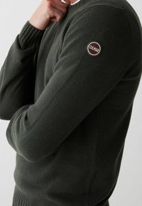 Tmavě zelený pletený svetr s žebrovanými manžetami a lemem, na levém rukávu je kulatá nášivka s logem Colmar. Klasický kulatý výstřih.