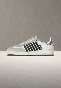 Baskets blanches avec des rayures noires, des accents en daim beige, un talon métallique argenté et le texte « DEAN » sur le côté.