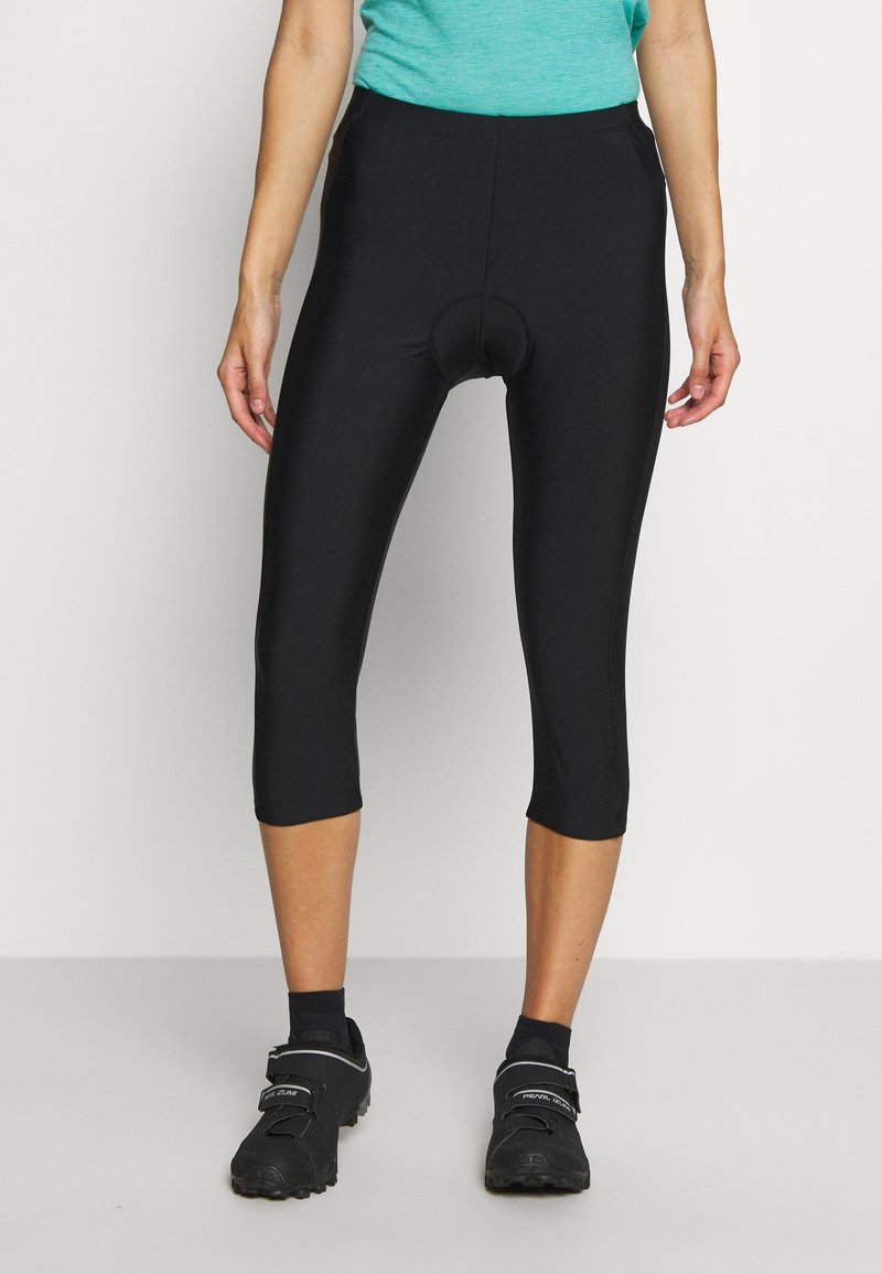 Femme portant un cuissard de cyclisme rembourré noir, des chaussures de cyclisme noires et un haut bleu sarcelle sur un fond uni.