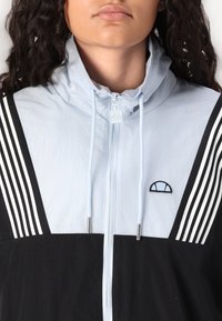 Lys blå og svart vindjakke med hette, med hvite striper, glidelås og brodert logo på brystet. Glatt tekstur, sporty design.
