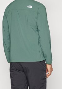 Zielona, lekka kurtka z długimi rękawami, z logo na plecach, gładką fakturą i prostym dołem, w zestawieniu z ciemnymi spodniami.