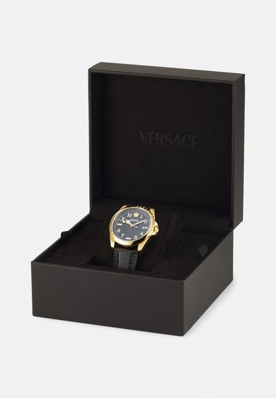 Aranyszínű óra fekete texturált bőrszíjjal és fekete számlappal, fehér számokkal és logóval, fekete dobozban bemutatva.