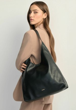 Kobieta w beżowym garniturze, niosąca dużą czarną skórzaną torebkę na ramię ze złotym logo, spoglądająca przez ramię na jednolitym tle.