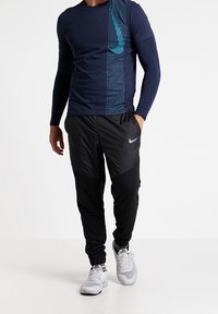 Camisa de manga larga azul marino con diseño de franjas en azul verdoso; pantalones de chándal negros, ajustados y con detalles grises; zapatillas deportivas blancas.