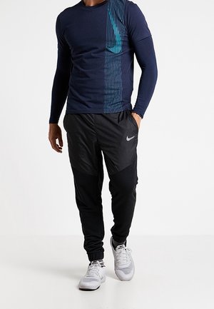 Camisa de manga larga azul marino con diseño de franjas en azul verdoso; pantalones de chándal negros, ajustados y con detalles grises; zapatillas deportivas blancas.