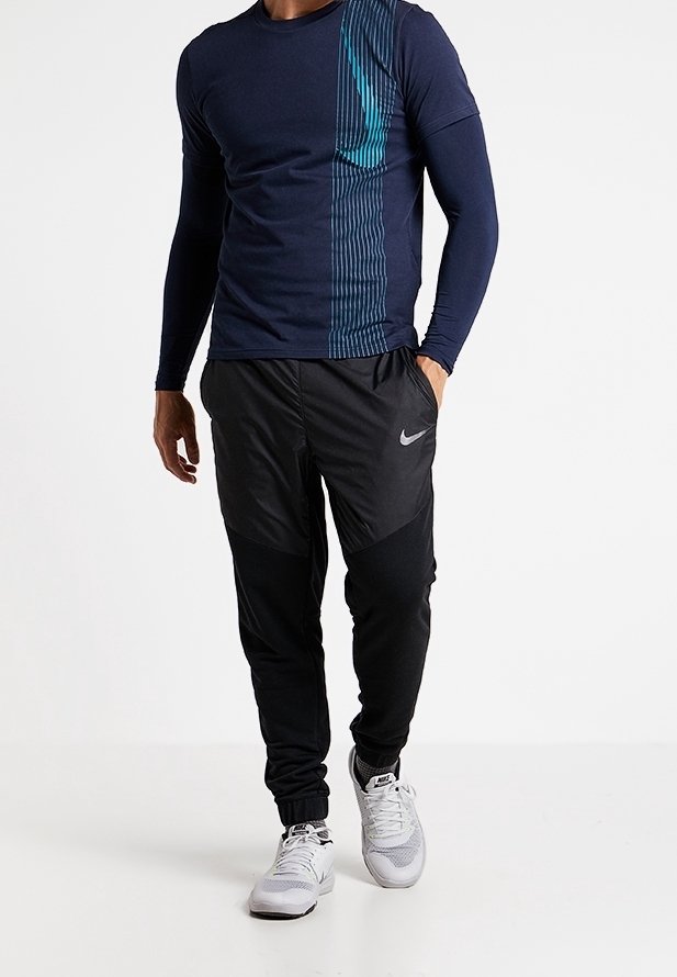 Camisa de manga larga azul marino con diseño de franjas en azul verdoso; pantalones de chándal negros, ajustados y con detalles grises; zapatillas deportivas blancas.