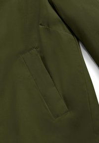 Giacca verde oliva con una texture liscia, tasca angolata e design minimalista, realizzata in tessuto leggero e resistente.