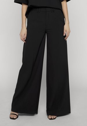 Schwarze, weit geschnittene Hosen, getragen von einer stehenden Person, kombiniert mit offenen schwarzen Sandalen vor einem einfarbigen hellen Hintergrund.