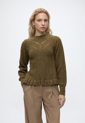 Jeune femme portant un pull vert olive texturé avec un ourlet à volants et un pantalon beige à jambes larges, debout devant un fond blanc uni.