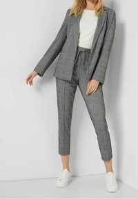 Femme portant un blazer à carreaux gris et un pantalon à cordon de serrage assorti, un haut blanc et des baskets blanches, debout contre un mur blanc.