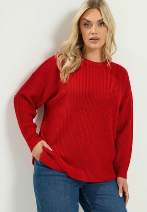 Lyshåret kvinde iført en løs rød strikket sweater og blå jeans, poserer med den ene hånd i lommen mod en ensfarvet hvid baggrund.