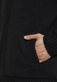 Cardigan negru din tricot, cu o textură ribbată și mansete, cu o mână plasată într-un buzunar lateral, evidențiind delicatețea materialului.