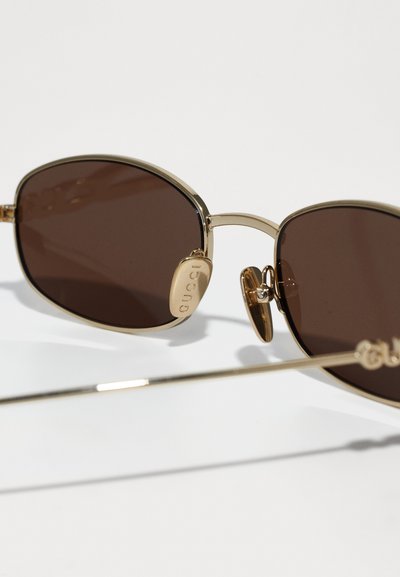 Gafas de sol de metal dorado con lentes marrones, forma ovalada, almohadillas nasales ajustables, con el logo de "Gucci" en el lateral.
