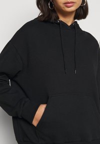 Femme portant un sweat à capuche noir avec poche avant et cordons, main reposant dans la poche, boucles d'oreilles anneaux visibles.