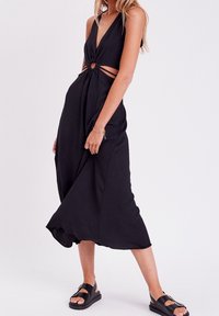 Robe noire sans manches avec un décolleté en V plongeant, des découpes et un nœud à la taille. Tissu léger avec un ourlet fluide et longueur cheville.