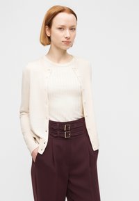 Ανοιχτό μπεζ ζακέτα με στρογγυλή λαιμόκοψη και κουμπιά μπροστά, φορεμένη πάνω από λευκό ριπ τοπ, συνδυασμένη με παντελόνια μπορντό ψηλόμεσα που διαθέτουν διπλές αγκράφες.