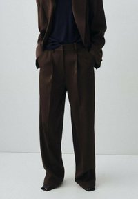 Pantalon marron taille haute avec des plis, jambes larges et une texture lisse. Associé à un haut sombre et des chaussures à bout ouvert noires.