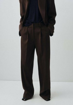 Pantalones marrones de cintura alta con pliegues, piernas anchas y una textura suave. Combinados con una parte superior oscura y zapatos negros de punta abierta.