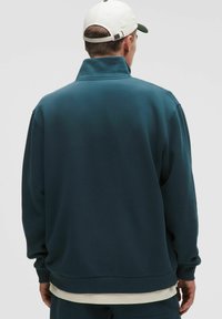 Tyrkysový pulóver so zvýšeným golierom, textúrovaná tkanina, spadnuté ramená a rebrované manžety. Nosený cez svetlú košeľu.