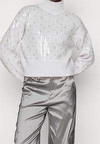 Pull blanc en maille avec un col montant, orné de sequins et d'un ourlet côtelé. Associé à un pantalon cargo argenté brillant.