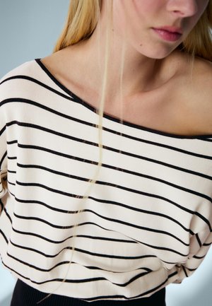 Femeie blondă purtând un top crem cu dungi orizontale negre, cu umerii goi, tăiat la gât și talie, pe un fundal deschis.