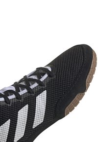 Svart idrottssko med vita ränder, svarta snören, texturerad mesh-övre och brun gummisula, vinkelrätt för att visa fram- och sidodetaljer.