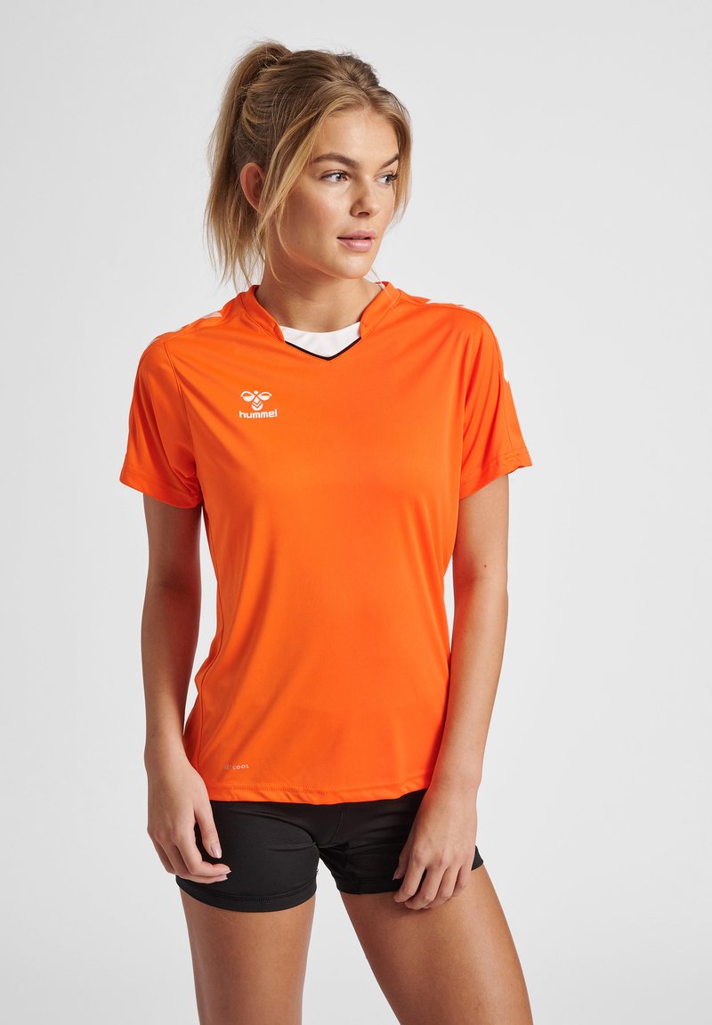 Femme aux cheveux blonds attachés en queue de cheval, portant un maillot de sport orange et un short noir, regardant sur le côté sur un fond clair uni.