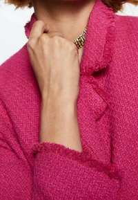 Femme portant une veste rose texturée avec des bords effilochés, tenant son poing près de son cou, avec un collier de perles multicolores visible.