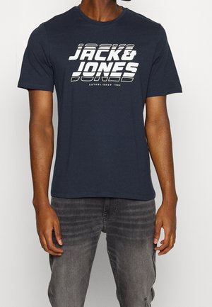Homme portant un t-shirt Jack & Jones bleu marine et un jean gris délavé, debout bras relâchés devant un fond blanc uni.
