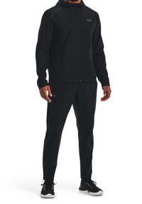 Musta urheilullinen verkkopuku, jossa on vetoketjullinen takki ja yhteensopivat housut. Kevyt kangas, istuva muotoilu ja minimaalinen brändäys.