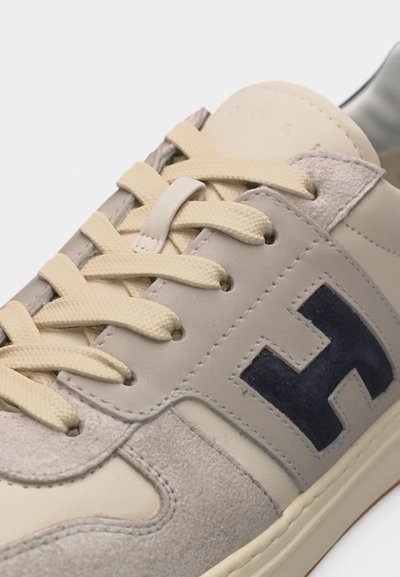Μπεζ αθλητικό παπούτσι με κρεμ κορδόνια, πάνελ από σουέτ και δέρμα, και ένα σκούρο μπλε λογότυπο "H" στην πλευρά, εμφανιζόμενο σε κοντινό πλάνο.