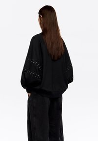 Sudadera negra con mangas abullonadas, con detalles de tachuelas en los brazos. Confeccionada con tela suave, diseñada con un corte holgado y puños acanalados.