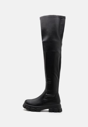 Botte haute en cuir noir avec semelle épaisse à crampons et finition lisse, conçue pour une couverture jusqu'au genou et un style robuste.