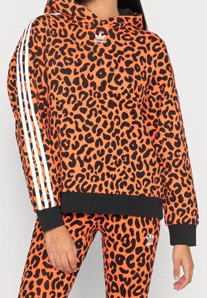 Femme portant un sweat à capuche Adidas imprimé léopard orange et noir et un legging assorti avec des détails à trois bandes blanches sur les manches et un logo sur la poitrine et la jambe.