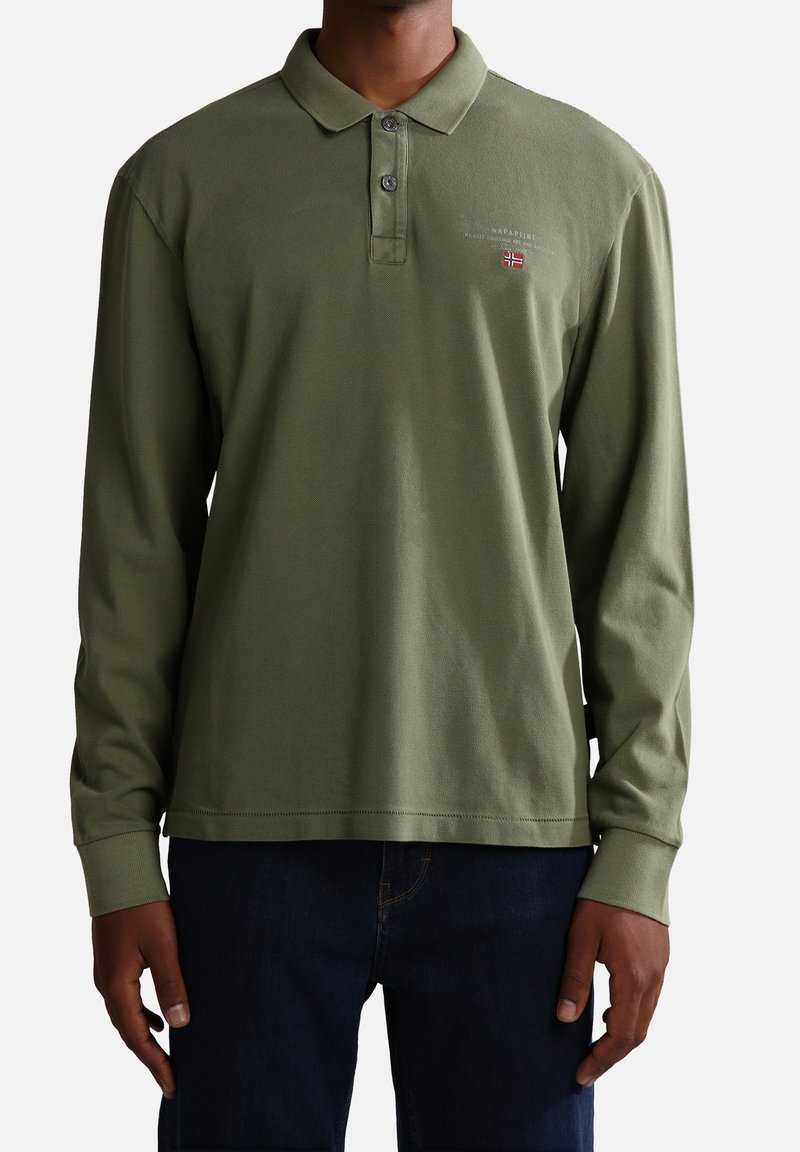 Homme portant un polo vert olive à manches longues avec patte de boutonnage et petit drapeau brodé sur la poitrine, associé à un jean bleu foncé.