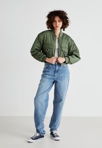 Chaqueta bomber verde oliva con dos bolsillos frontales, herrajes plateados y puños acanalados, combinada con unos vaqueros holgados azul claro y zapatillas azul marino.