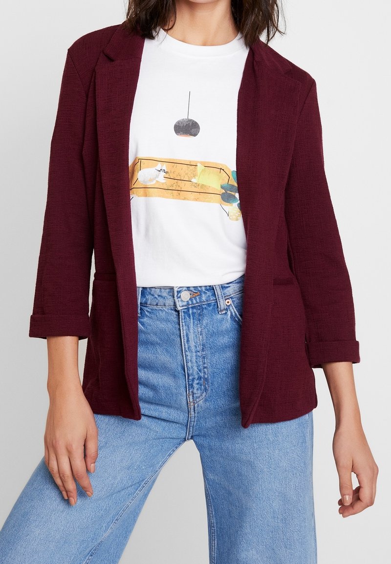 Burgundský, texturovaný blazer s otevřeným předním designem, dvěma předními kapsami, ohrnutými rukávy, vrstvený přes bílé tričko s potiskem a světle modré džíny.