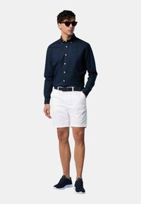 Camicia a maniche lunghe navy con bottoni, pantaloni corti bianchi e sneakers navy. Gli occhiali da sole e la cintura intrecciata completano l'outfit.