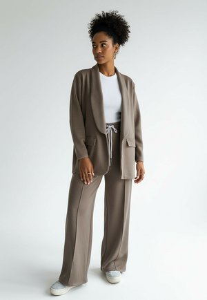 Zestaw do salonu w jasnobrązowym odcieniu, składający się z długiej kurtki z kołnierzem szalowym, dwoma przednimi kieszeniami oraz pasujących szerokich spodni z troczkiem. Pod spodem biały top.