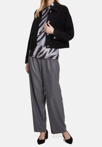 Žena v černé zkrácené zapínací bundě, zebra potiskem na topu, šedých širokých kalhotách a černých lodičkách stojí na bílém pozadí.