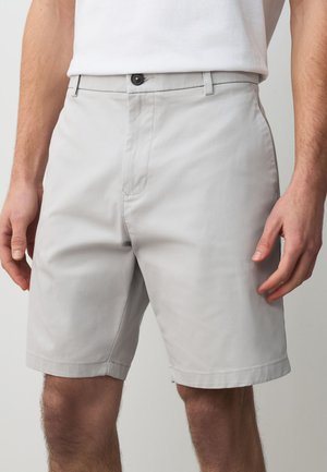 Uomo che indossa pantaloncini su misura grigio chiaro e una camicia bianca, con le mani rilassate ai lati, mostrato dalla vita alle ginocchia.