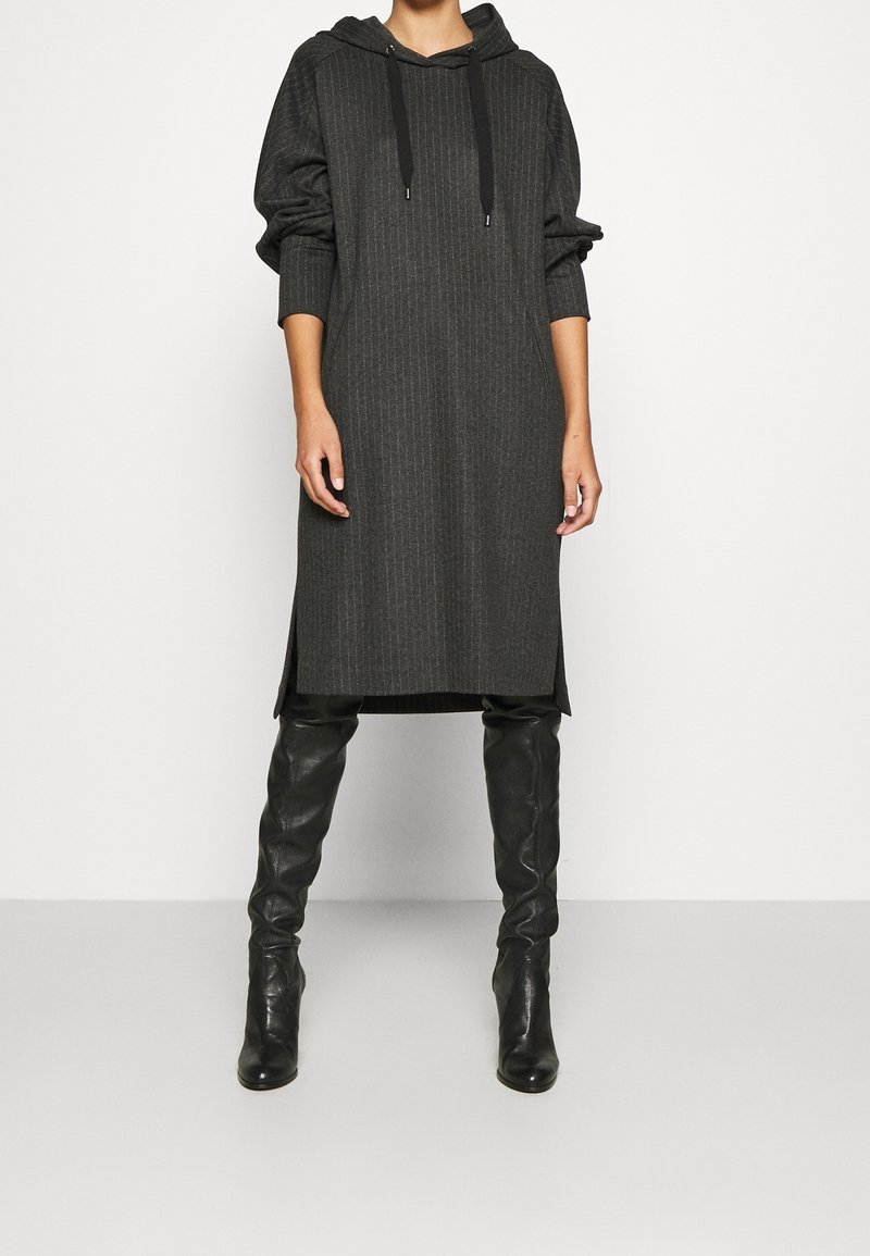 Personne portant une robe à capuche à rayures fines gris foncé avec des manches longues, associée à des bottes en cuir noires montantes sur un fond uni.