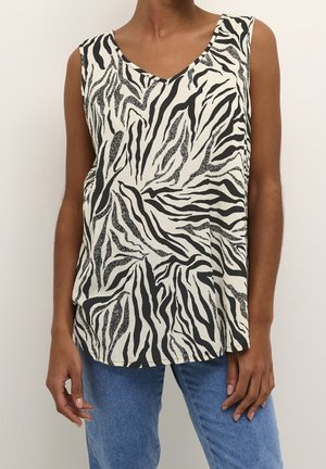 Personne portant un haut sans manches avec un motif zèbre noir et blanc et un jean bleu, debout devant un fond clair uni.