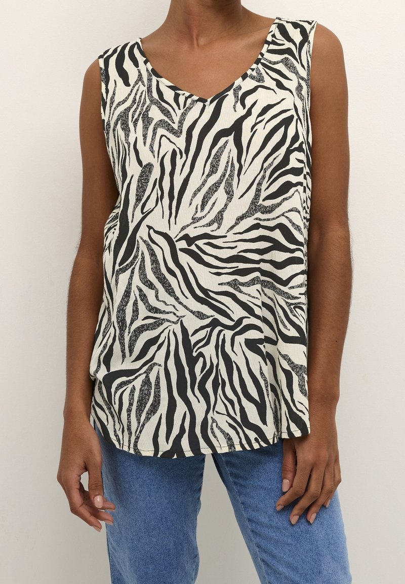 Personne portant un haut sans manches avec un motif zèbre noir et blanc et un jean bleu, debout devant un fond clair uni.